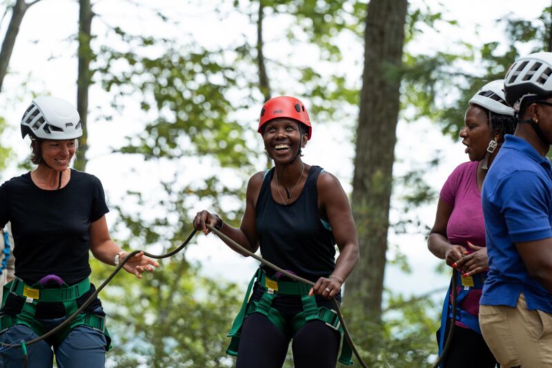 The image shows a group of people wearing helmets and harnesses, likely preparing for or participating in an outdoor activity like a ropes course or climbing. They are smiling and appear to be enjoying themselves in a wooded area. The focus is on the woman in the center with a red helmet, who is holding a rope and laughing.
